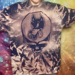 May include: A tie-dye t-shirt with a large graphic featuring a wolf-like creature and a skull design. The shirt has a bleached, mottled pattern in shades of brown and black. The graphic is in a dark blue color.