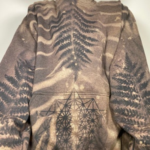 Puede incluir: Una sudadera con capucha marrón con un diseño de helecho negro y un patrón geométrico en la parte delantera. La sudadera tiene un aspecto desgastado, con áreas descoloridas y una sensación vintage.