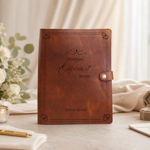 Peut inclure: Un classeur en cuir marron avec une fermeture à pression argentée, gravé avec le texte "Wedding Officiant Binder" et "Taylor Marie", orné de motifs de papillons dans les coins et d'une bordure rectangulaire sur la couverture avant.