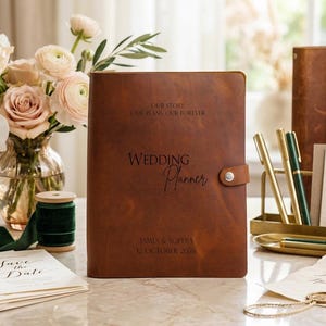 May include: A brown leather wedding planner with a snap closure. The cover is engraved with the text: OUR STORY, OUR PLANS, OUR FOREVER, WEDDING PLANNER, JAMES & SOPHIA, 12 OCTOBER 2026. The planner is positioned upright on a flat surface.