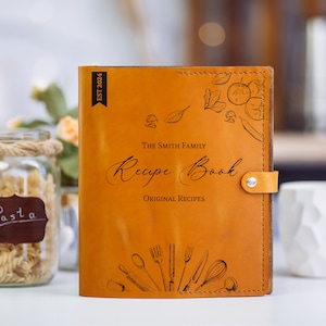 May include: A tan leather recipe book with a snap closure. The cover features the text "The Smith Family Recipe Book," "Original Recipes," and "EST 2024," alongside black line art illustrations of various kitchen utensils and fresh vegetables.