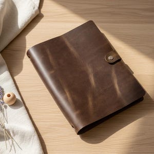 May include: A dark brown leather journal with a button clasp. The journal is closed and placed on a wooden surface. A white cloth, a wooden bead, and lavender sprigs are nearby.