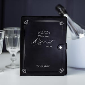 May include: A black leather-like binder with a snap closure, featuring the white printed text "WEDDING Officiant BINDER" and "TAYLOR MARIE" with butterfly corner accents. The binder is positioned between two clear glass goblets and a white ice bucket.