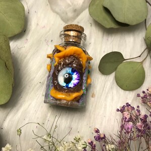 May include: A small glass bottle with a cork stopper is decorated with purple, green and orange polymer clay. A large blue and purple eye is embedded in the clay.