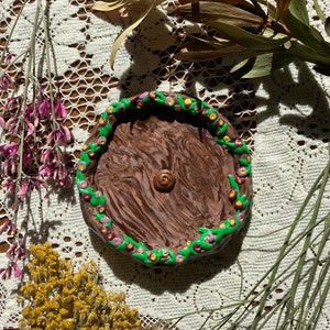 Puede incluir: Un plato de arcilla marrón y rosa con un diseño floral verde y morado alrededor del borde. El plato tiene un diseño de espiral en el centro.