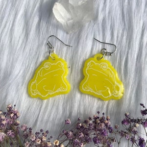 Puede incluir: Un par de pendientes de rana amarillos con contornos blancos. Los pendientes están hechos de resina y tienen ganchos plateados.