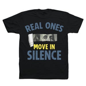 May include: Black t-shirt with the text "REAL ONES MOVE IN SILENCE" in blue and yellow. The design includes a torn paper effect revealing a portion of a US $100 bill.