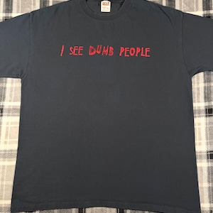 May include: Dark gray t-shirt with the red text "I SEE DUMB PEOPLE". The shirt has a crew neck and short sleeves. The shirt is made of a soft material.