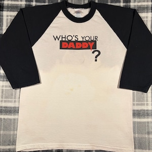 May include: Cream-colored raglan shirt with black sleeves. The shirt features the text "WHO'S YOUR DADDY?" in black and red, with a question mark. The shirt is made of a soft material.