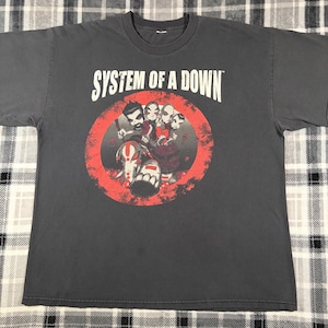 May include: Dark gray t-shirt featuring the band name "SYSTEM OF A DOWN" in white block letters above a red circular graphic. The graphic depicts several stylized figures with red and white accents.