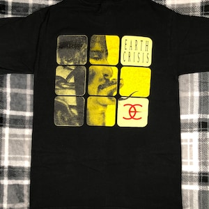 May include: Black t-shirt with a yellow and black graphic of a person's face in a grid pattern. The text "EARTH CRISIS" is printed in white above the graphic. The graphic also includes a red "EC" logo.