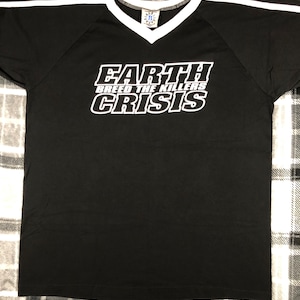 May include: Black t-shirt with white stripes on the shoulders and a white V-neck collar. The shirt features the text "EARTH BREED THE KILLERS CRISIS" in a bold, white, stylized font. The shirt is made of a soft material.