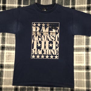 May include: A navy blue t-shirt with a white graphic print of "Rage Against The Machine" with stars around the text.