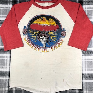 May include: Cream-colored raglan shirt with red sleeves. The front features a circular graphic with a skull, roses, and the words "Grateful Dead". The graphic includes a phoenix and lightning bolts.