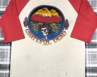 May include: Cream-colored raglan shirt with red sleeves. The front features a circular graphic with a skull, roses, and the words "Grateful Dead". The graphic includes a phoenix and lightning bolts.