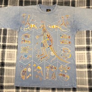 May include: Light blue t-shirt featuring a colorful kangaroo and other Australian animal illustrations in a unique, artistic style. The design includes a variety of animals and geometric patterns, creating a vibrant and eye-catching design.