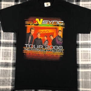 May include: Black t-shirt with a graphic of the band *NSYNC on their "No Strings Attached" tour in 2000. The graphic features the band members in front of a red, orange, and yellow background with the text "TOUR 2000" and "NO STRINGS ATTACHED" in white.