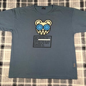 May include: A slate blue short-sleeve t-shirt featuring a graphic of a stylized bear head with large blue eyes and sharp teeth. Below the bear is a black rectangular box containing text about a test specimen and Radiohead copyright info.
