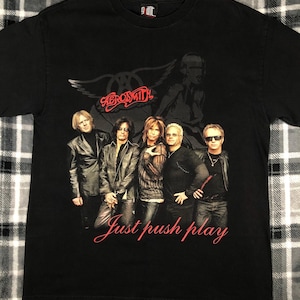 May include: Black t-shirt with a graphic of the band Aerosmith. The band members are pictured in front of a winged symbol. The text "Just push play" is printed below the image.