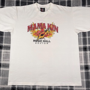 May include: White t-shirt featuring the text "MAMA KIN" in red, with a flame design and a musical instrument graphic. Below the text is "MUSIC HALL BOSTON". The shirt is made of a soft material.