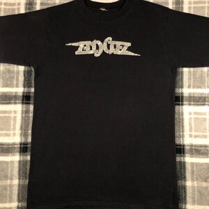 May include: Black t-shirt with the word "EDGEZ" in a silver, glittery font across the chest. The text is stylized with a lightning bolt design. The shirt is made of a soft material and has a crew neck.