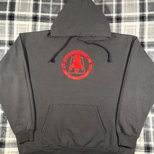 May include: A gray hooded sweatshirt with a red rocket ship logo inside a circle on the front. The sweatshirt has a front pocket and a drawstring hood. The background is a plaid pattern.
