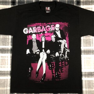 May include: Black t-shirt featuring the band Garbage with a pink halftone design. The band members are depicted in a stylized illustration. The band's name is in white block letters above the image. The shirt has a crew neck.
