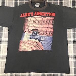 May include: Dark gray t-shirt with the text "JANE'S ADDICTION" in red block letters. The shirt features a graphic of a person in bed under a patterned blanket. The shirt has a gray collar.