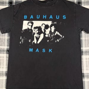 May include: Dark gray t-shirt featuring the band Bauhaus with the word "BAUHAUS" in blue above a black and white image of the band members. The word "MASK" is printed in blue below the image.