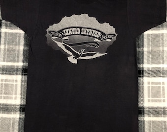 Puede incluir: Camiseta negra con un gráfico blanco de un pájaro volando frente a una nube con el texto "LYNYRD SKYNYRD" en una pancarta.