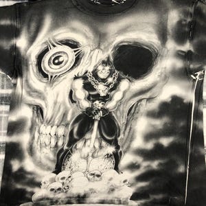 May include: Black and white graphic t-shirt featuring a muscular figure with a skull-shaped head, holding a large weapon, standing on a cloud of skulls. The background is a large skull. The shirt has a crew neck.