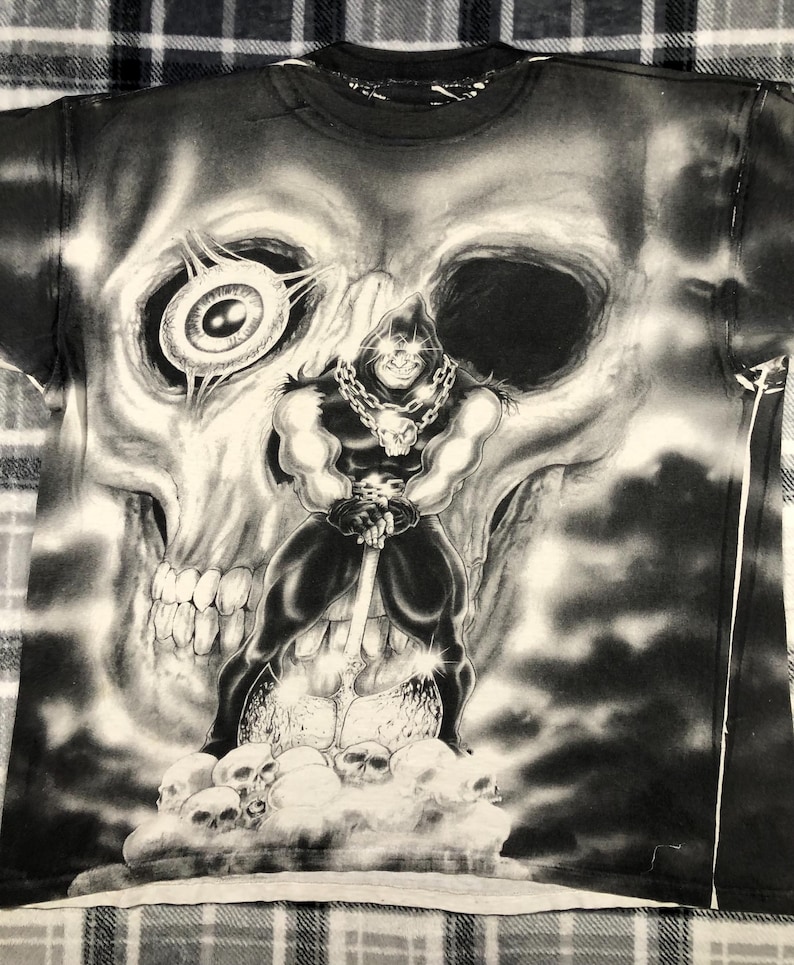May include: Black and white graphic t-shirt featuring a muscular figure with a skull-shaped head, holding a large weapon, standing on a cloud of skulls. The background is a large skull. The shirt has a crew neck.