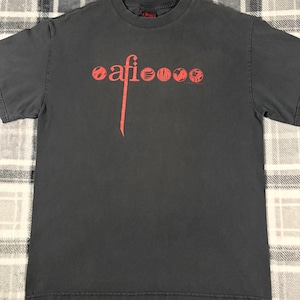 May include: Dark gray t-shirt with the word "afi" in red, stylized with a vertical line and several circular designs. The shirt is short-sleeved and appears to be made of cotton. The background is a plaid pattern.