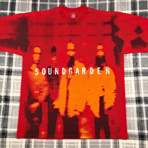 May include: A red short-sleeve t-shirt featuring a graphic print of four figures in orange and dark red tones. The word SOUNDGARDEN is printed in white across the center of the chest. The shirt has a crew neckline and a small globe icon at the bottom.