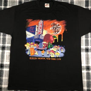 May include: Black t-shirt with a graphic of the Allman Brothers Band performing at the Beacon Theater in New York City. The graphic features a red apple, a truck, and mushrooms. The text "Where it all Begins" and "Join the Peach Corp" are also included in the graphic.