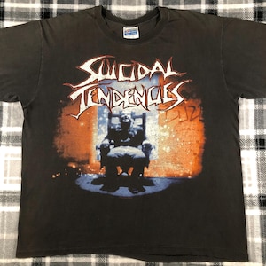 May include: Black t-shirt with a graphic print of the band "Suicidal Tendencies" in red and white lettering. The graphic features a person sitting in a chair in front of a brick wall with orange and blue accents.