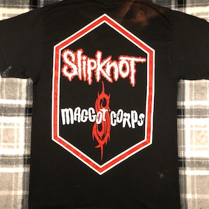 May include: Black t-shirt with a red and white hexagonal logo featuring the band name "Slipknot" and the text "Maggot Corps".