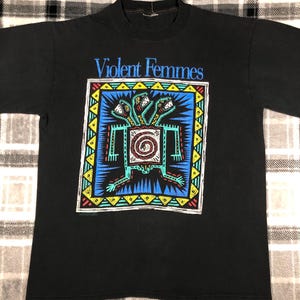 May include: Black t-shirt featuring the band name "Violent Femmes" in blue above a colorful graphic. The graphic includes a central figure with a spiral design, surrounded by a blue and yellow border, and three-headed creature.