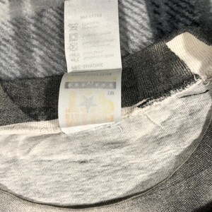 May include: Close-up of a gray and white t-shirt with a tag attached. The tag is white with black text and symbols. The t-shirt is made of a soft, textured fabric. The tag appears to have washing instructions.