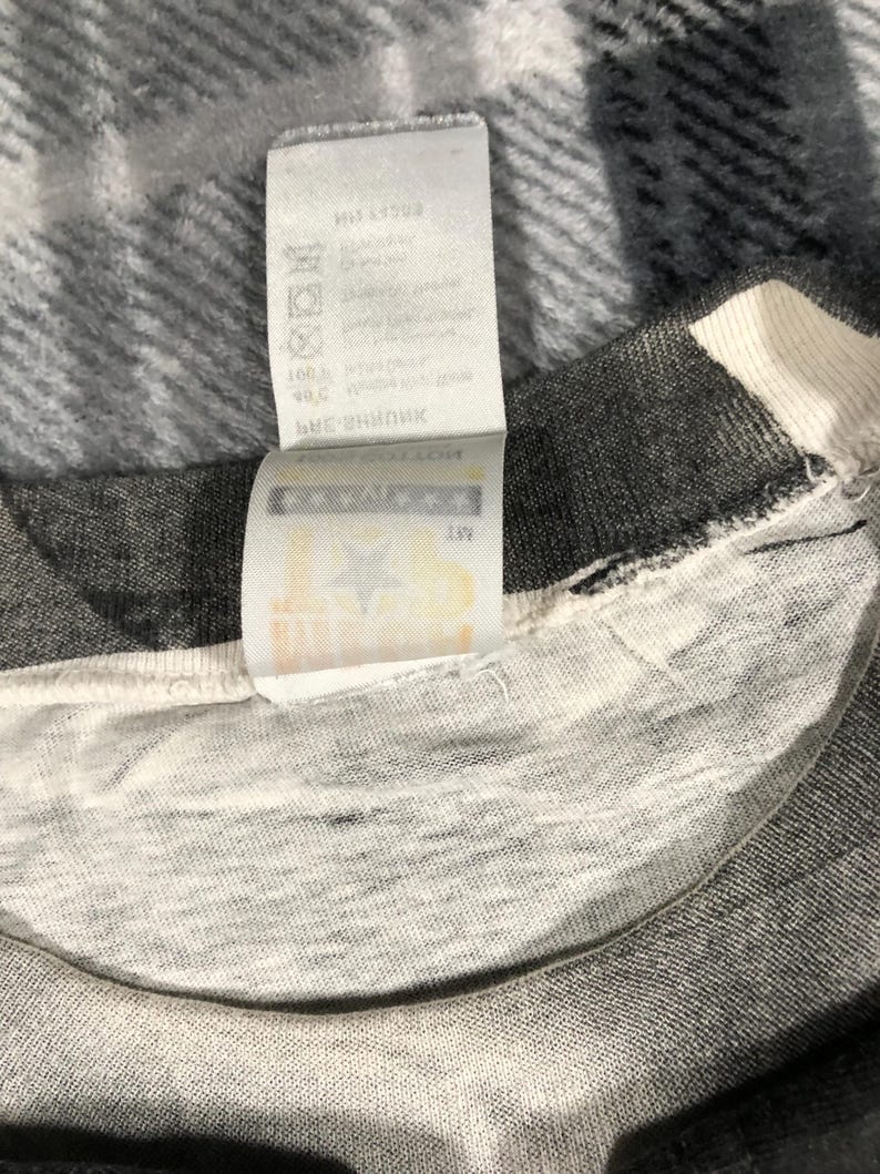 May include: Close-up of a gray and white t-shirt with a tag attached. The tag is white with black text and symbols. The t-shirt is made of a soft, textured fabric. The tag appears to have washing instructions.