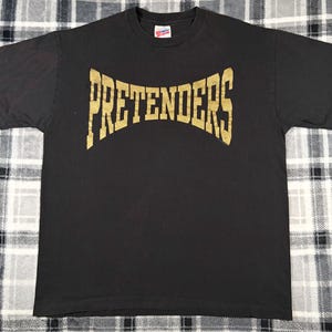 May include: Black t-shirt with the word "PRETENDERS" in gold, arched lettering across the chest. The shirt is made of a soft material and has a crew neck. The shirt is laid flat on a checkered surface.