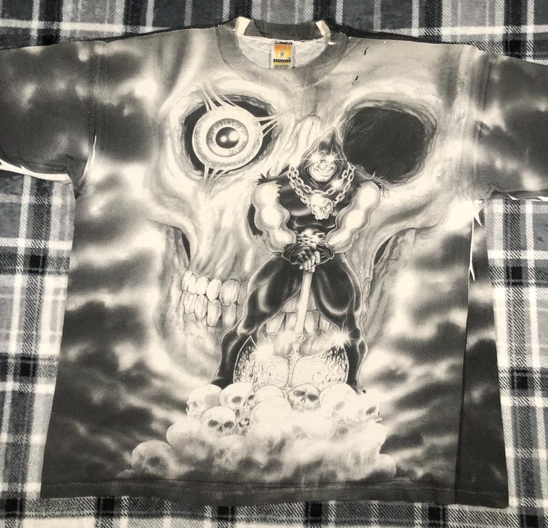 May include: A gray t-shirt featuring a black and white graphic of a muscular figure holding a sword, standing in front of a large skull. The design includes multiple skulls and a chain around the figure's neck. The shirt has a crew neck.