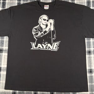 May include: A black short-sleeve t-shirt featuring a white graphic print of a person wearing sunglasses and singing into a microphone. The name LAYNE is printed in large white capital letters below the graphic. The shirt has a crew neckline.