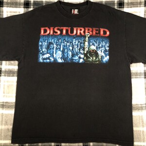 May include: Black t-shirt featuring the band name "DISTURBED" in red letters above a blue and white graphic of a crowd. The shirt is a crew neck and made of a soft material.