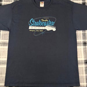 May include: A navy blue short-sleeve t-shirt featuring a graphic of a white electric guitar headstock. The text reads "The Original Fender Stratocaster" in blue and "Real gone... Since 1954!" in white, accented by thin blue lines.
