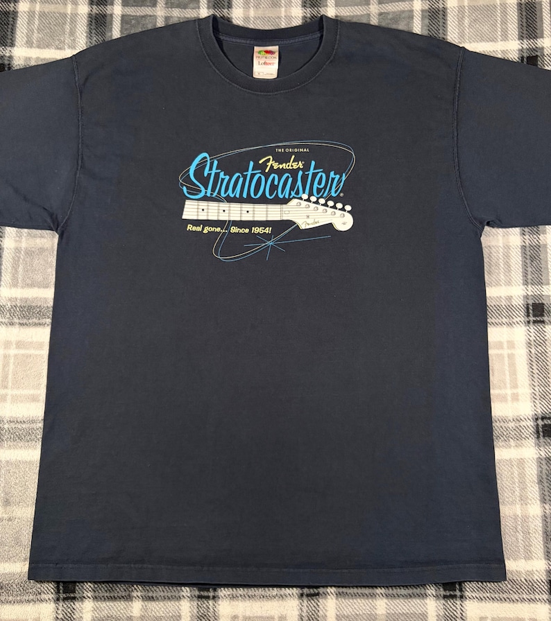 May include: A navy blue short-sleeve t-shirt featuring a graphic of a white electric guitar headstock. The text reads "The Original Fender Stratocaster" in blue and "Real gone... Since 1954!" in white, accented by thin blue lines.