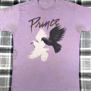 May include: A light purple t-shirt featuring the word "Prince" in a stylized purple font. Below the text are two doves, one white and one black, with red beaks. The shirt is a crew neck style.