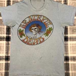 May include: A light blue t-shirt with a vintage Grateful Dead logo featuring a skull with flowers and the words "Grateful Dead" in a circular design.