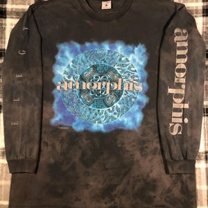 May include: Dark gray long-sleeve shirt featuring a blue and teal circular graphic with the band name "Amorphis". The shirt has the word "Elegy" printed vertically on one sleeve and "Amorphis" on the other.