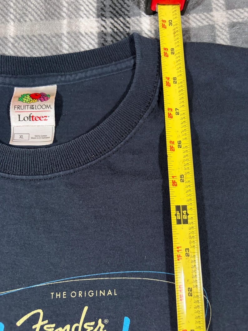 May include: A dark navy blue t-shirt with a crew neckline and a Fruit of the Loom Lofteez label indicating size XL. A yellow measuring tape is placed vertically along the fabric, showing a length measurement extending past 30 inches.
